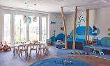 Ein heller Raum in der Kita kinderzimmer Tienrade, ein blauer Wal und weitere Unterwassertiere wurden an die Wand gemalt, ein Bereich zum spielen ist mit einem Teppich versehen und auf dem Holzfußboden steht ein kleiner Tisch mit Stühlen daran