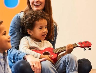 Eine Frau hat ein Kind auf dem Schoß sitzen, welches eine kleine Gitarre in der Hand hält, ein anderes Kind sitzt daneben