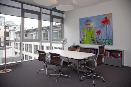 Ein Büro mit einem weißen Tisch und vier Stühlen daran, eine Fensterfront und ein Regal über dem ein Bild hängt