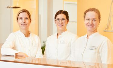 Drei Frauen in jeweils einem weißen Kittel, stehen hinter einem Empfangstresen und lächeln in die Kamera