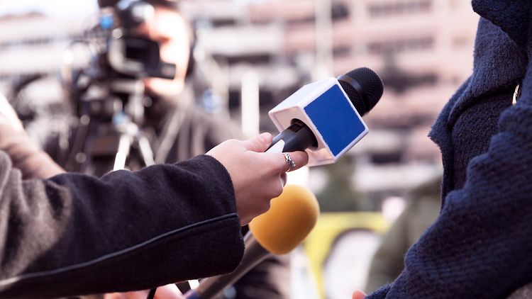  Einer Person werden vor einer Kamera im Hintergrund zwei Mikrofone für ein Interview hingehalten.
