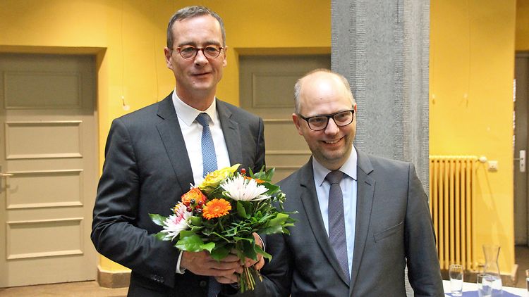  Zwei Männer im Anzug. Der linke erhält vom rechten Mann Blumen überreicht.