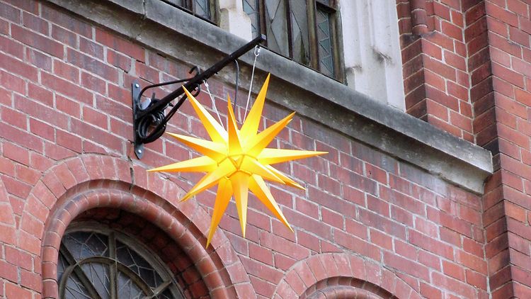  Ein gelber dreidimensionaler vielzackiger Stern hängt an einem Seil über mehreren Fenstern eines rot geklinkerten Gebäudes.