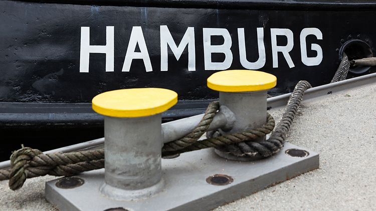  Die Schrift "HAMBURG" steht auf einem schwarzen Schiffsbug. Im Vordergrund ist der Kai und die Tampen, mit dem das Schiff an diesem fixiert ist.