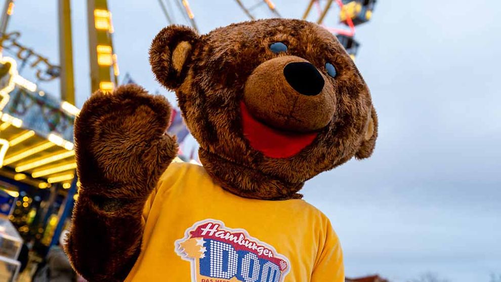  Ein Mensch trägt ein Kostüm, dass aussieht, wie ein brauner flauschiger Teddybär. Er trägt ein gelbes T-Shirt mit der Aufschrift Hamburger DOM.
