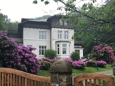  Weiße Villa mit Holzverkleidung, im Vordergrund lila Rhododendren