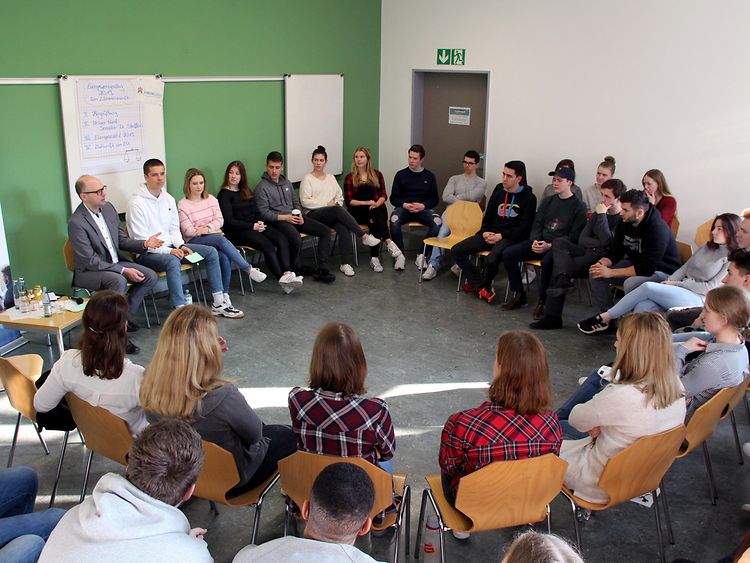  Ein Stuhlkreis in einem Klassenraum aus Schülerinnen und Schülern und einem Mann in Anzug.