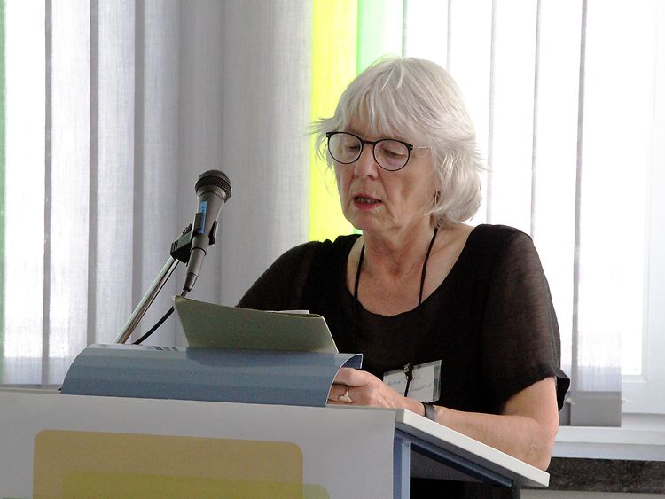  Eine ältere Frau mit grauen Haaren steht hinter einem Rednerpult und spricht.