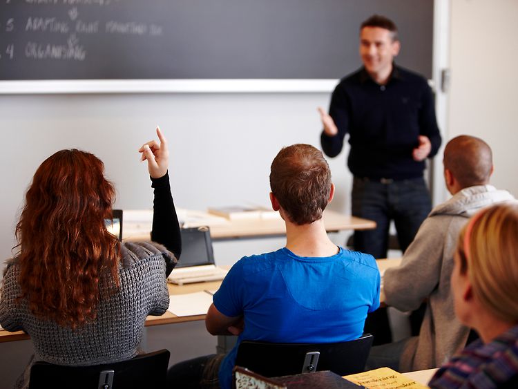  Ein Lehrer steht vor einer Tafel und nimmt eine Schülerin dran, die ihren Finger hebt. Drei weitere Schüler sitzen neben ihr.