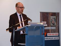  Ein Mann mit Anzug, Krawatte und lichtem Haar steht hinter einem Rednerpult. Rechts neben ihm steht ein Banner: "Jobs bei der Hamburger Justiz"