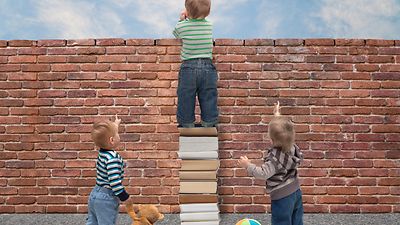  Ein Kind steht vor einer Mauer auf einem Bücherstapel, zwei andere kleine Kinder zeigen nach oben und flankieren links und rechts.