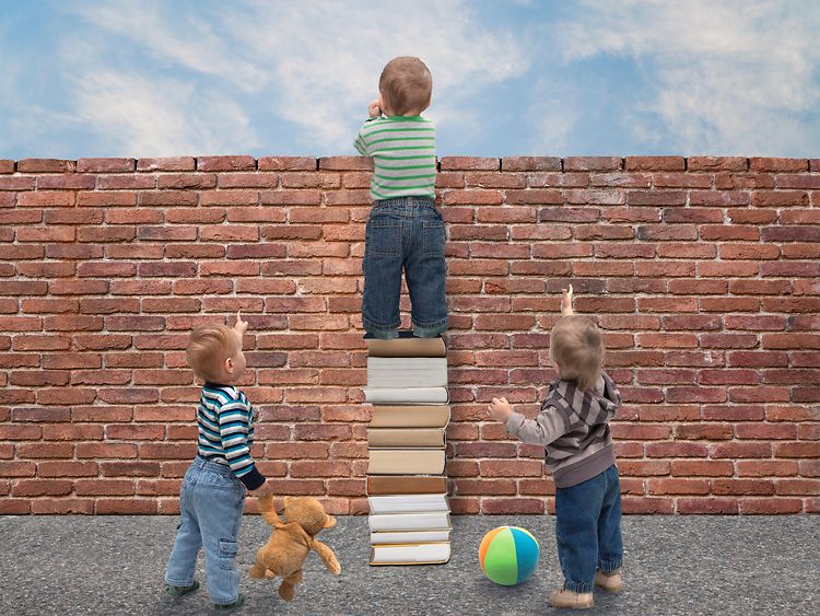  Ein Kind steht vor einer Mauer auf einem Bücherstapel, zwei andere kleine Kinder zeigen nach oben und flankieren links und rechts.
