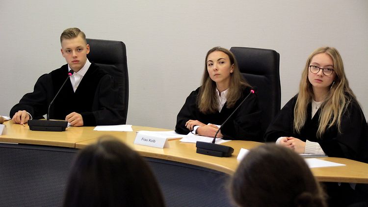  Drei Schülerinnen und Schüler sitzen in schwarz-weißer Richterrobe vor den Klägern.