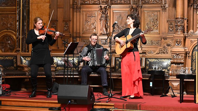 Das Trio der Band "A Mekhaye" spielt auf der Bühne im Rathaus. Der Gesang wird durch eine Violine, ein Akkordeon und eine Gitarre begleitet. 