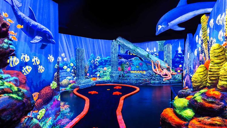 Minigolf in einer Unterwasserwelt mit Fischen und Korallenriffen, Pizza im Bistro und Menschen, die sich durch Laserstrahlen bewegen.
