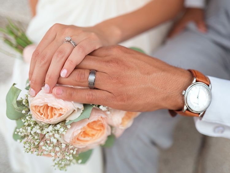  Ein Mann und eine Frau halten ihre Hände über einen Blumenstrauß. Beide tragen einen silbernen Ring, der Mann trägt eine Uhr mit braunem Lederband.