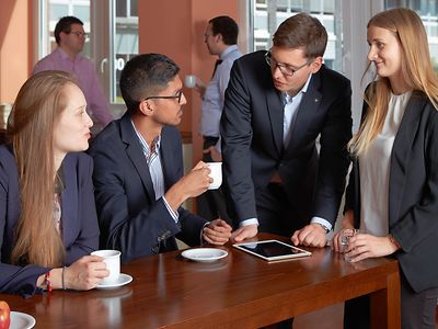  vier Personen in Businesskleidung an der Cateringinsel mit Kaffee und Tablet im Gespräch vertieft.