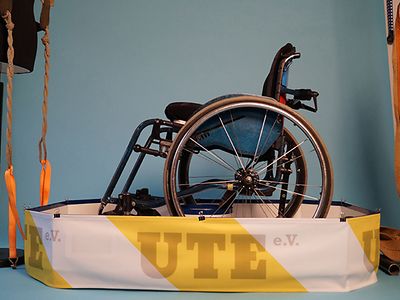  ein Rollstuhl mit einer Trageaufhängung, mit der zwei Personen ihn tragen können