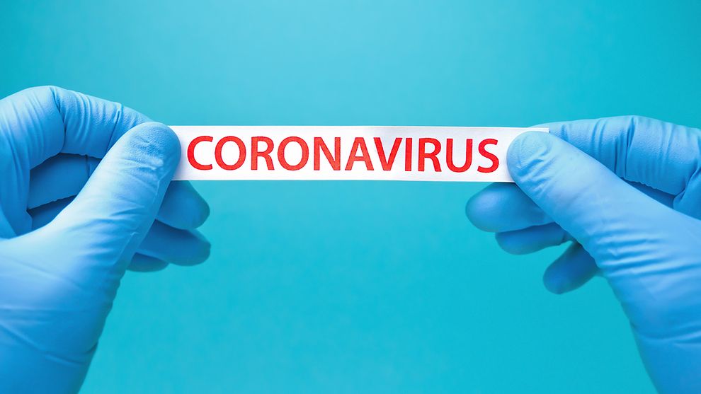 Zwei Hände in Einmalhandschuhen halten ein Zettel, auf dem "Coronavirus" steht.