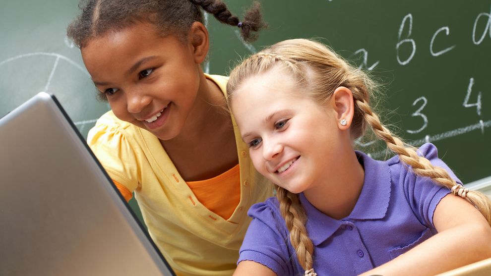 Zwei Mädchen mit Lehrerin am Laptop vor einer Tafel im Klassenraum