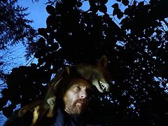  Mann mit einem Fuchs über dem Kopf steht im dunklem Wald.w
