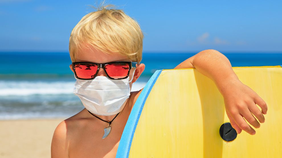 Junge mit Maske lehnt am Strand vor dem Meer an seinen Surfboard