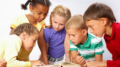  Fünf Kinder beugen sich gemeinsam über ein aufgeschlagenes Buch auf einem Tisch (Symbolbild)