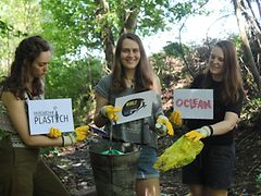  Teresa Mohr, Jana Wohlgethan und Alina Bendlin beim Cleanup im Wald. Sie halten Schilder mit den Logos der Initiative Plastich, des World-Cleanup-Days und von Oclean in die Kamera.