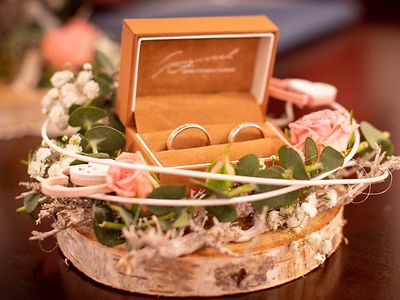  Auf dem Bild liegen zwei Ringe in einer Schmuckschatulle. Umgeben wird die Schatulle von Blumen, die auf einer Holzplatte angerichtet sind.