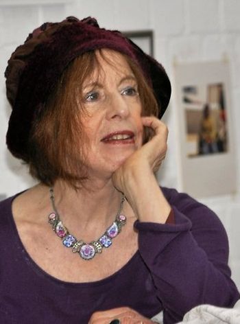 Die Malerin Karin Witte sitzt, das Gesicht in die Hand gestützt und mit einem lilafarbenen Hut geschmückt in ihrem Hamburger Atelier.
