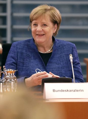 Bundeskanzlerin Angela Merkel, gekleidet in ein marineblaues Kostüm, sitzt an einem Tisch und lächelt in Richtung Kamera. Vor ihr ein Mikrofon und ein Schild, auf dem "Bundeskanzlerin" zu lesen ist.