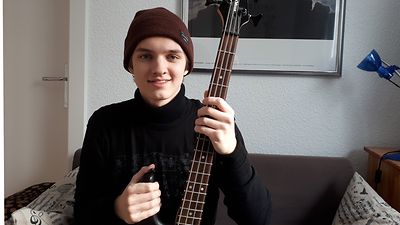  ein junger Mann mit Mütze sitzt auf dem Sofa mit einem E-Bass in der Hand
