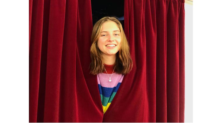 Eine junge Frau steckt den blonden Kopf durch einen roten Theatervorhang