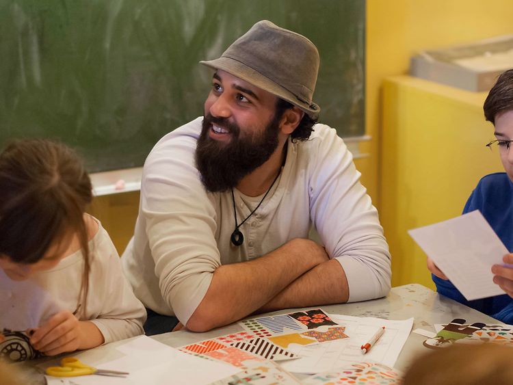  ein junger Mann mit Vollbart und modischem Hut zwischen einem Mädchen und einem Jungen im Kunstunterricht