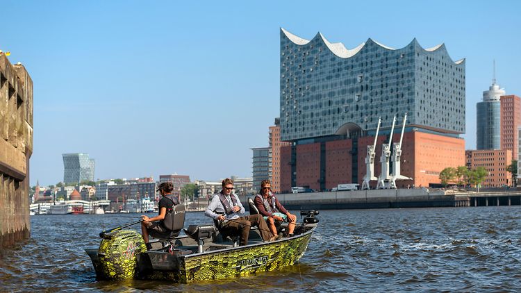  Drei Angler sitzen in einem kleinen Boot auf der Elbe vor der Elbphilharmonie und angeln. 