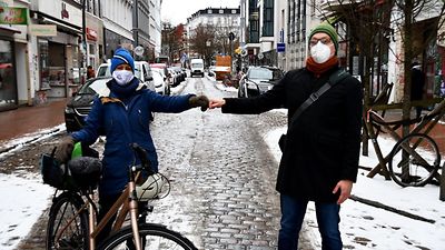  Zwei Personen mit Maske, davon eine mit Fahrrad, stehen auf einer Straße und geben sich die Faust.