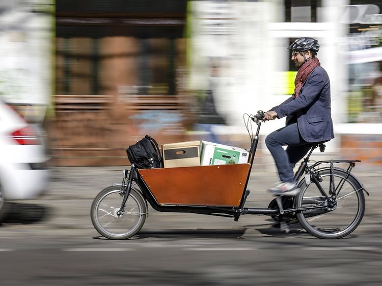  Ein Mann mit Helm fährt auf einem Lastenrad mit Kartons im Behälter.