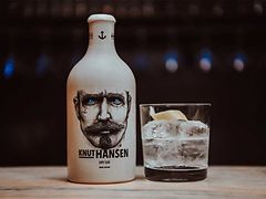  Auf dem Bild sind eine weiße Flasche mit dem Motiv des Gesichts eines Mannes und ein mit einer Flüssigkeit gefülltes Glas zu sehen. Auf der Flasche steht Knut Hansen Dry Gin.