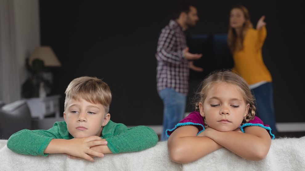 Zwei Kinder auf einer Couch, im Hintergrund streiten zwei Erwachsene.