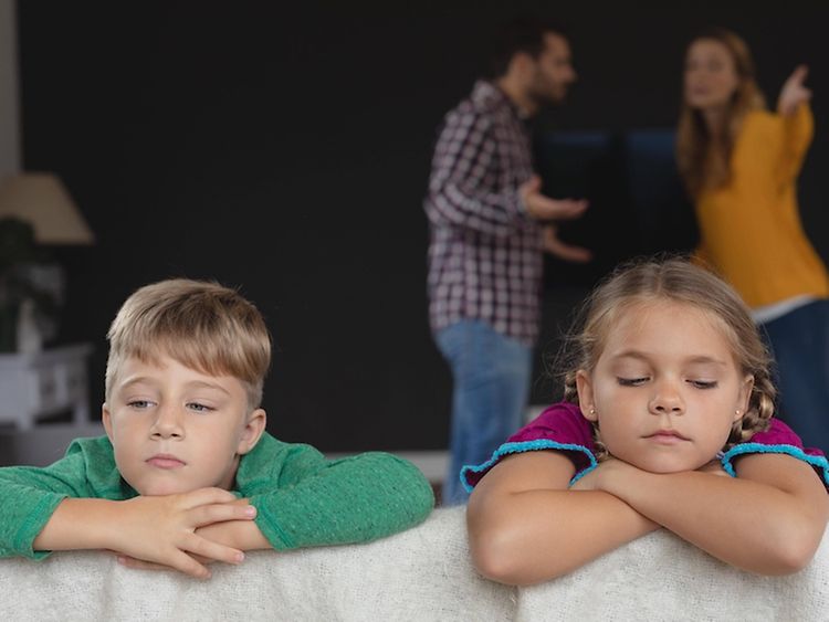  Zwei Kinder auf einer Couch, im Hintergrund streiten zwei Erwachsene.