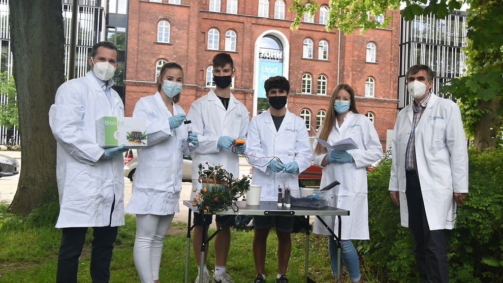 Zwei Lehrpersonen und vier Schüler stehen im weißen Kittel vor der Technischen Universität. Vor Ihnen steht ein Tisch mit Experimentier-Utensilien.