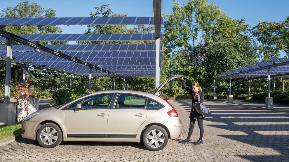 Eine Frau betätigt die Kofferraumtür eines Autos auf einem Parkplatz, welcher mit Photovoltaik-Anlagen überspannt ist.