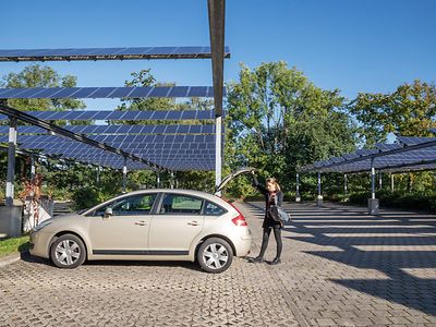  Eine Frau betätigt die Kofferraumtür eines Autos auf einem Parkplatz, welcher mit Photovoltaik-Anlagen überspannt ist.
