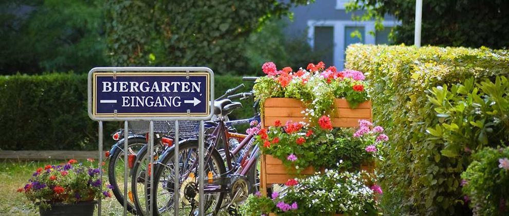  Ein blaues Schild, mit der Aufrischt "Biergarten Eingang" am Eingang eines Biergartens. Im Umfeld des Schildes befinden sich Fahrräder, bepflanzte Blumenkästen sowie Bäume und Hecken. 