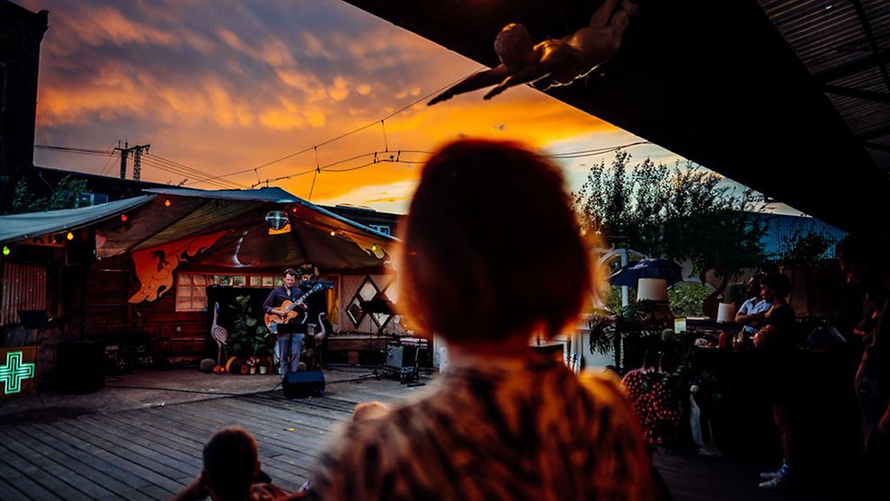Eine Frau bei einer OpenAir Veranstaltung in der Ensemble Resonanz. Zu sehen ist ein Sänger mit Gitarre und orangener Himmel im Abendrot.