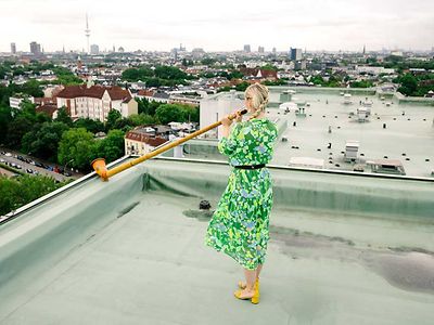  Eine Frau in einem grüngemusterten Kleid steht mit einem Horn auf dem Dach und blickt über Hamburg.