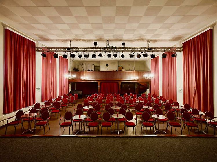  Der Saal des kleinen hoftheaters mit roten Stühlen und Holztischen. An den Wänden hängen rote Vorhänge.