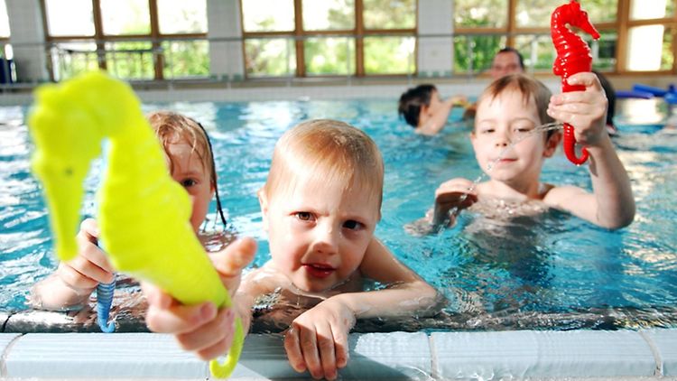  Drei Kinder im Schwimmbecken eines Hallenbades. Der Junge in der Mitte hält ein gelbes, aufblasbares Seepferdchen in die Kamera. Ein Junge rechts neben ihm hält ein rotes Seepferdchen hoch.