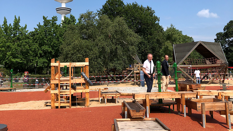  Zwei Personen stehen auf einem Spielplatz mit rotem Boden und Holzegräten.