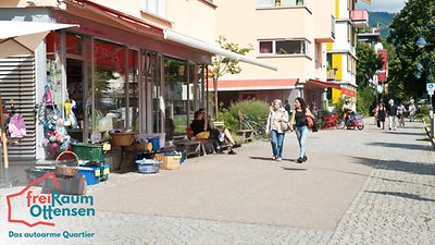  Straße im Freiburger Stadtteil Vauban, auf der sich ausschließlich Fußgänger und Radfahrer bewegen. Rechter Bildrand: Ein Laden hat seine Ware unter einer Markise ausgebreitet.